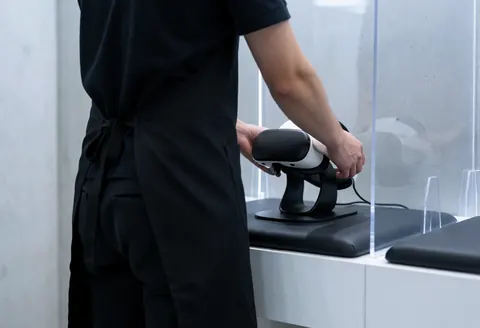 Un membre du personnel place un casque de réalité virtuelle sur une station de charge, derrière une barrière transparente, dans un espace avec murs en béton et sol rembourré.
