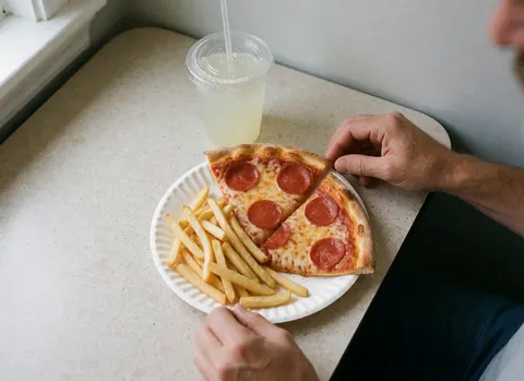 Des mains saisissant une part de pizza pepperoni et des frites sur une assiette en papier, accompagnées d'un verre de limonade avec paille sur une table.