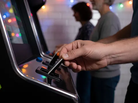 Une main adulte insère une carte dans la fente d'une borne arcade noire avec des lumières de fête floues en arrière-plan et des murs carrelés blancs.