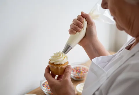 Une personne âgée portant un tablier blanc applique du glaçage sur un cupcake dans un atelier de cuisine professionnel.