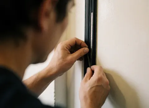 Mains du technicien utilisant un outil pour ajuster le joint magnétique d'une porte de réfrigérateur sur fond blanc chaud, vue rapprochée des détails de la réparation.