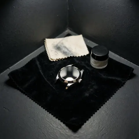 Boîtier de montre brillant posé sur un tissu noir, accompagné d'un chiffon et d'un pot de composé de polissage dans un coin aux murs gris.