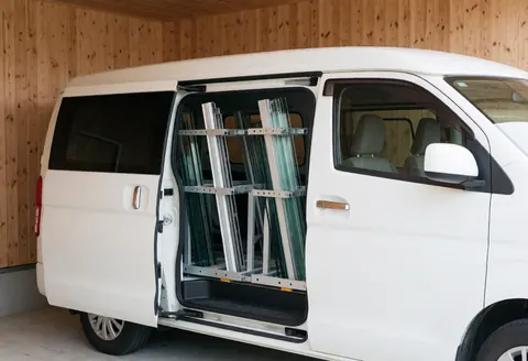 Fourgonnette blanche stationnée dans un garage aux murs en bois clair, porte ouverte révélant des étagères chargées de panneaux de verre et de profilés en aluminium, baignée par la lumière naturelle du matin.