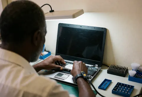 Technicien en tenue antistatique remplace un disque dur sur un ordinateur portable ouvert, avec outils et lumière de travail sur une table propre.