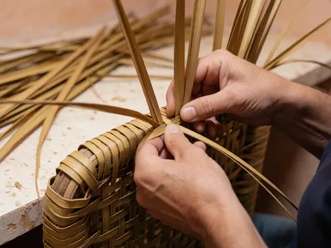 Mains d'artisan tissant des feuilles de palmier sur un cadre de chaise traditionnel dans un atelier avec des murs en terre et des outils visibles.