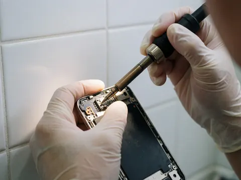 Technicien en gants réparant une prise jack 3, 5 mm sur un smartphone avec un fer à souder, fond de carreaux blancs, détails de circuit et outil visible.