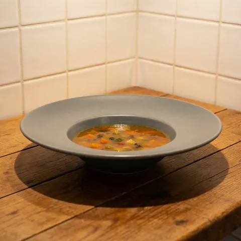 Soupe aux légumes servie dans un bol en céramique grise mate posé sur une table en bois rustique, avec des carreaux blancs au fond et une lumière chaleureuse.