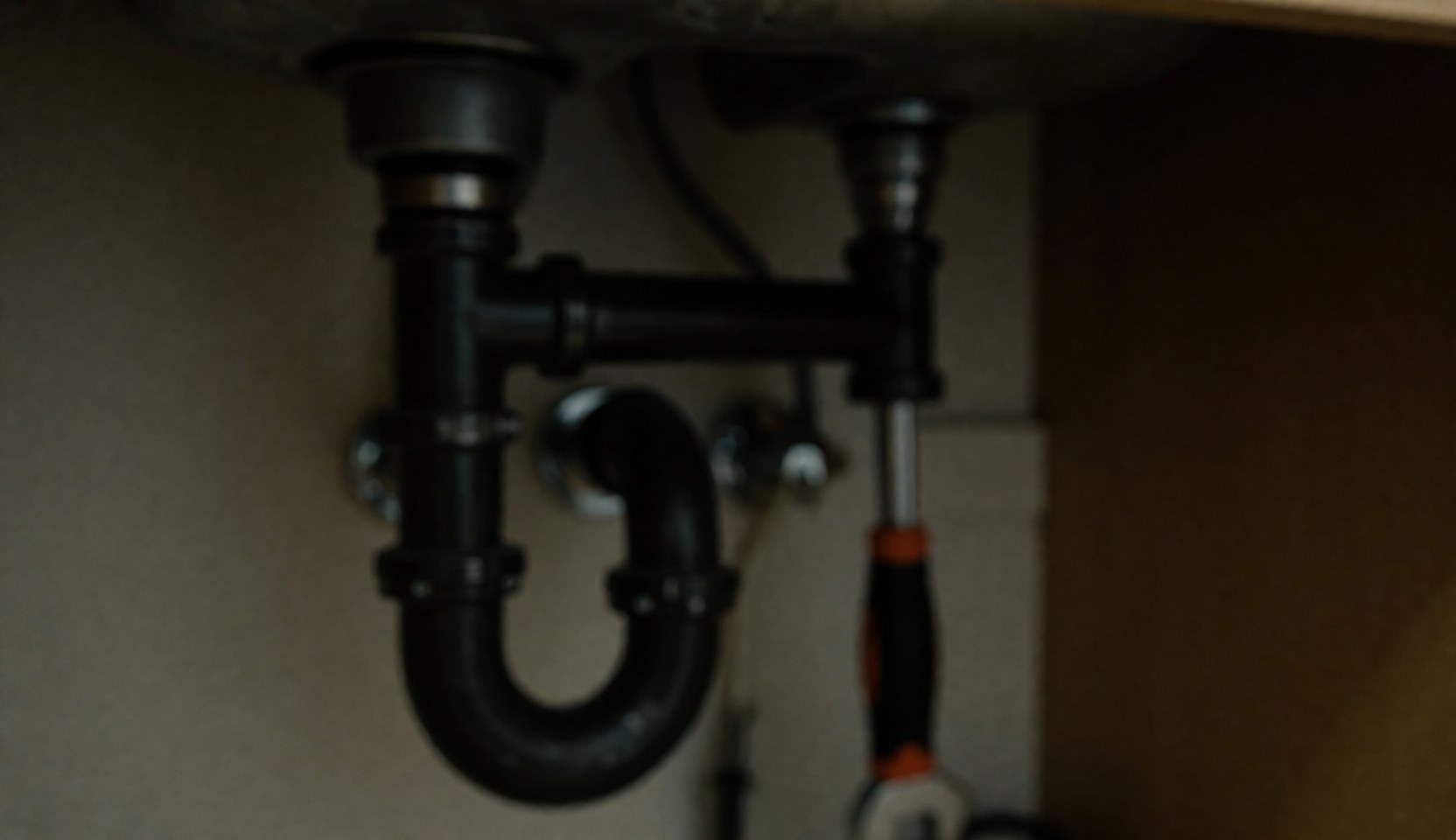 Vue rapprochée des tuyaux sous un évier de cuisine, mettant en évidence un siphon en P et une clé à pipe posée sur un fond flou pour un texte.