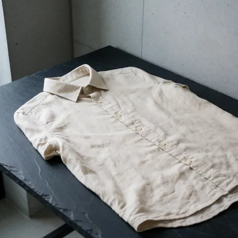 Chemise en lin beige avec boutons posée sur une table en pierre sombre, murs en béton, lumière naturelle, texture visible, design minimaliste.
