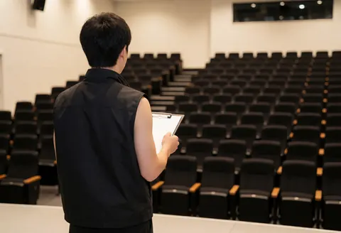 Un technicien portant un gilet noir vérifie l'installation d'une salle de conférence vide avec des rangées de chaises et un tableau blanc.
