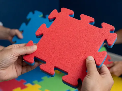 Des mains adultes tiennent une pièce de puzzle rouge à bordure bleue sur fond coloré dans un parc d'attractions.