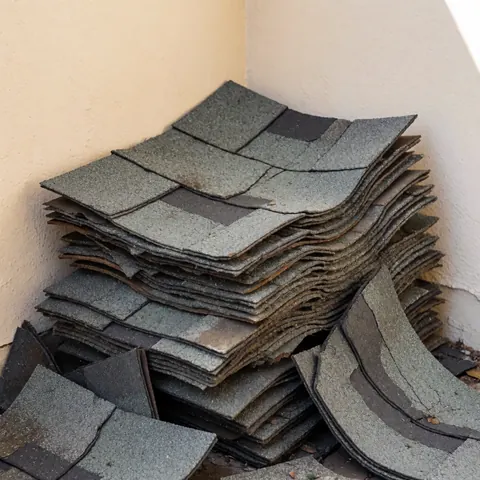 Empilement de tuiles d'asphalte usées et fissurées dans un coin, révélant texture granuleuse et couches irrégulières sur fond beige.