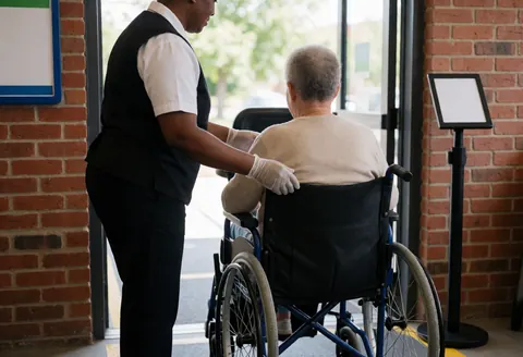 Un employé de parc à thème avec des gants aide une personne âgée en fauteuil roulant près d'une entrée avec un mur en briques.