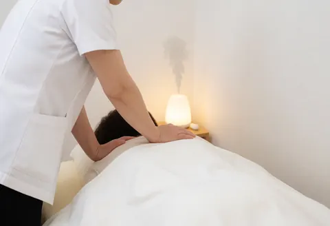 Un thérapeute en uniforme blanc effectue un massage dorsal sur un client couvert d'une serviette blanche, dans une pièce aux murs blancs avec un diffuseur émettant de la vapeur.