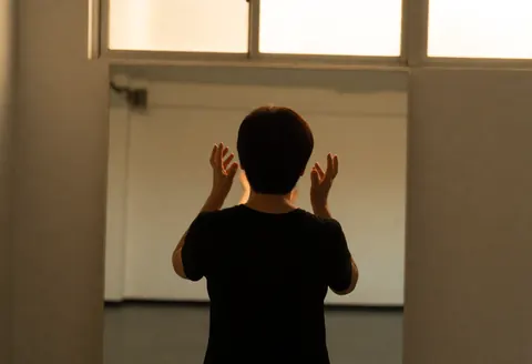 Une personne vue de dos effectue des exercices de théâtre face à un grand miroir dans une salle d'essai baignée de lumière naturelle.
