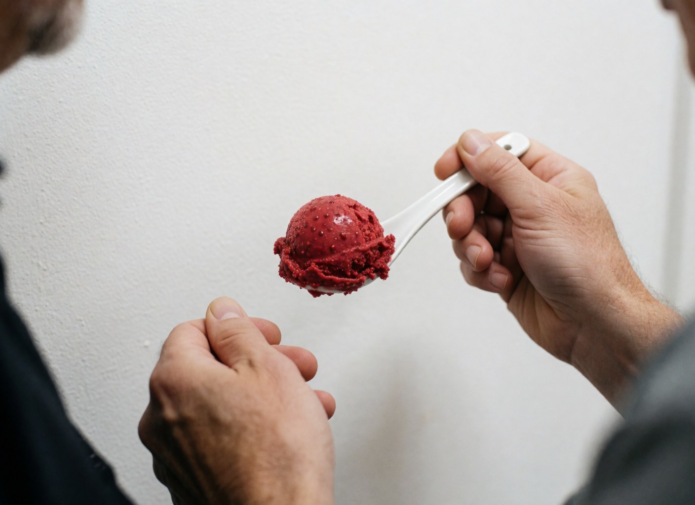 Glace aux framboises avec des graines, servie dans une petite cuillère blanche tenue par des mains sur un fond blanc.