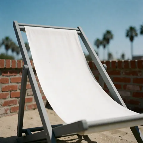Chaise longue blanche avec cadre gris sur le sable, devant un mur en briques rouges et des palmiers flous sous un ciel bleu.