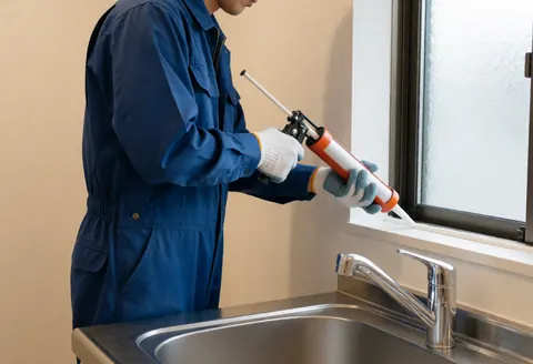 Un ouvrier en combinaison bleue applique un joint blanc sur le cadre d'une fenêtre au-dessus d'un évier en acier inoxydable dans une cuisine aux murs clairs.
