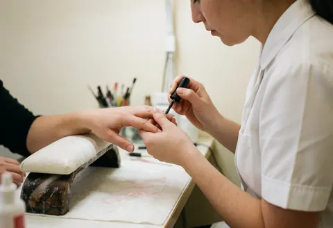 Une technicienne applique avec précision du vernis noir sur les ongles d'une cliente. Le bras repose sur un repose-pieds rembouronné dans un environnement propre et bien éclairé.