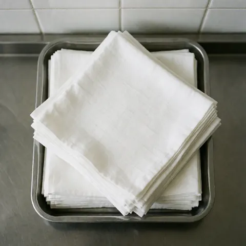 Des compresses stériles et propres empilées sur un plateau en acier inoxydable, posé sur une surface métallique polie, avec des murs de carreaux blancs en arrière-plan.