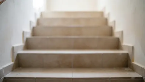 Vue d'escaliers intérieurs avec un sol en carreaux de couleur beige clair, des murs blancs et un effet de flou sur les éléments environnants, créant un fond neutre adapté aux textes commerciaux.