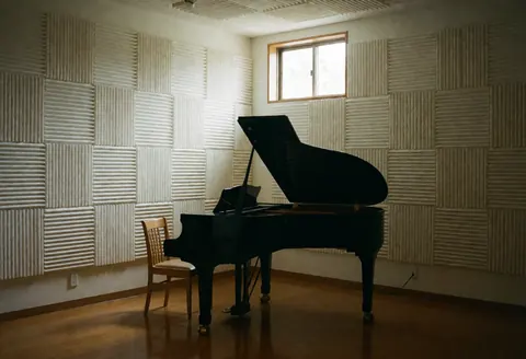 Salle de musique vide équipée d'un piano à queue noir, d'une chaise en bois et de panneaux acoustiques aux murs, baignée de lumière naturelle provenant d'une fenêtre haute.