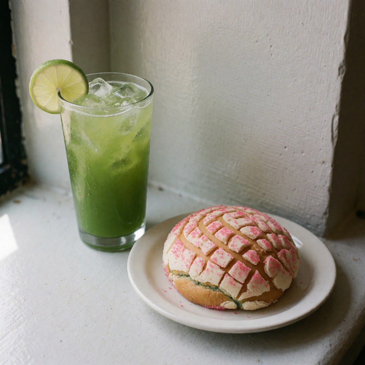 Verrée haute contenant une boisson verte glacée et des tranches de citron vert, accompagnée d'une coquille de sucre rose sur une assiette blanche près d'une fenêtre.