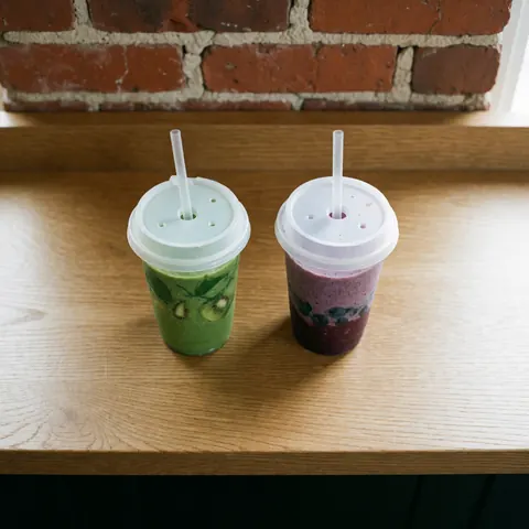 Deux boissons fraîches, l'une verte au kiwi et l'autre violette aux fruits, servies dans des verres sur une table en bois clair devant un mur de briques rouges.