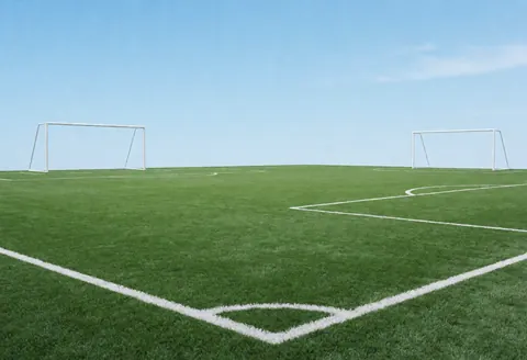 Vue d'un terrain de sport extérieur avec pelouse synthétique verte, lignes blanches délimitant la zone de jeu et deux buts blancs aux extrémités.