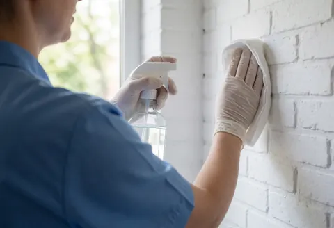 Un thérapeute en gants blancs nettoie un mur de briques blanches avec un chiffon et un spray désinfectant, éclairé par la lumière naturelle d'une fenêtre.
