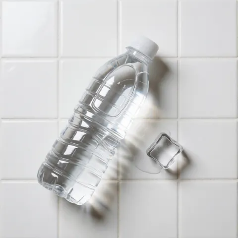 Bouteille d'eau transparente avec bouchon blanc et un glaçon posés sur des carreaux blancs, éclairage lumineux et propre, scène minimaliste pour hôtel ou auberge.
