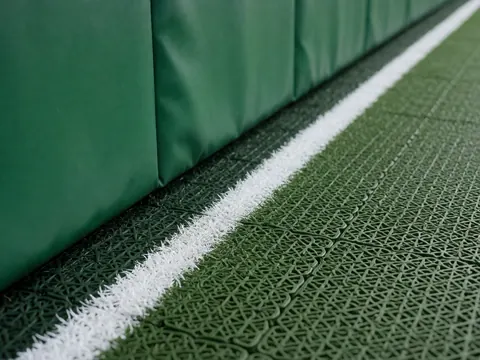 Gros plan détaillé du coin où le mur vert avec coutures rejoint le sol en gazon synthétique aux lignes blanches marquées, idéal pour un club sportif.