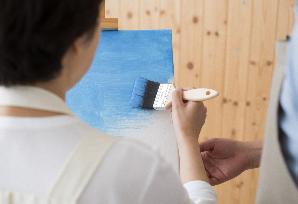 Une personne adulte peint sur une toile bleue avec un pinceau tandis qu'une main guide son bras dans un atelier aux murs en bois clair.