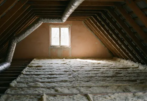 Plafond en construction avec isolation en laine de verre entre poutres, gaines métalliques et cloisons en plâtre rose éclairées par une fenêtre.