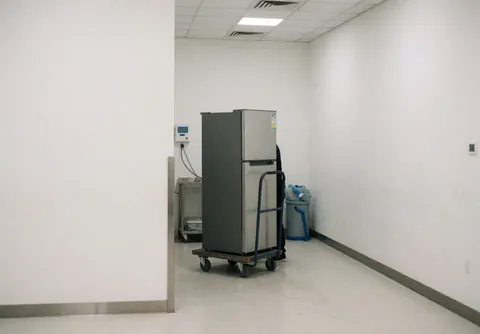 Un réfrigérateur à deux portes posé sur un chariot manuel au sein d'un atelier de réparation aux murs blancs et au sol propre.