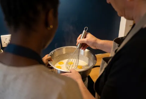 Un instructeur guide un étudiant lors du mélange d'œufs dans un bol en cuisine, avec des ustensiles visibles.
