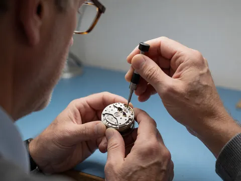 Technicien ajuste le balancier d'un mouvement de montre à l'aide d'un outil spécialisé sur une surface bleue dans un atelier de réparation.