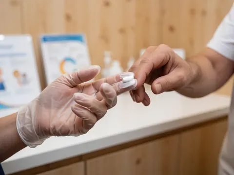 Gros plan d'une main portant un gant médical qui applique une crème sur un doigt, avec un fond en bois clair et un éclairage professionnel mettant en valeur la texture du produit.