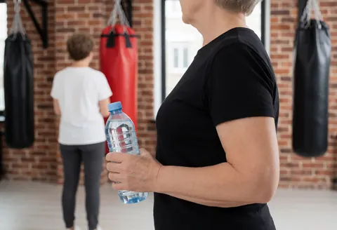 Une femme adulte tient une bouteille d'eau dans un gymnase avec des sacs de boxe et un mur en briques en arrière-plan.
