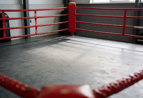 Ring de boxe vide aux cordes rouges sur sol en béton avec murs gris, idéal pour l'entraînement dans un environnement industriel.