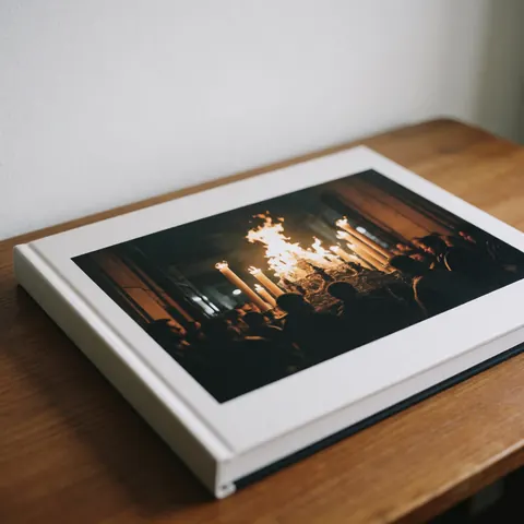 Album photo professionnel ouvert sur une table en bois, révélant une procession avec des bougies allumées et des vêtements sombres, éclairé par une lumière naturelle diffuse.