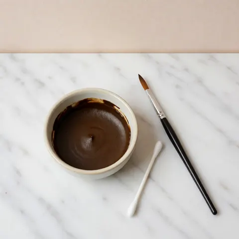 Récipient en céramique contenant du henné pour sourcils, accompagné d'un pinceau et d'un coton-tige posés sur une surface en marbre blanc, prêt pour une application professionnelle.