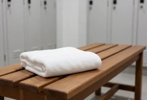 Une serviette blanche soigneusement pliée repose sur un banc en bois clair dans un vestiaire aux casiers blancs flous, baigné d'une lumière vive et d'une ambiance propre.