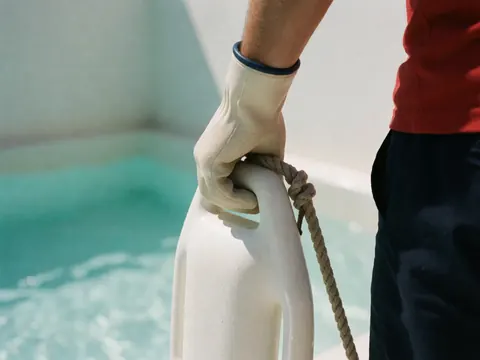 Gant de protection tenant une bouée blanche avec corde épaisse près d'une piscine turquoise ensoleillée.