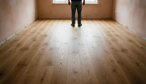 Un professionnel observe la pose d'un plancher en bois neuf dans une pièce vide aux murs enduits, baignée de lumière naturelle.