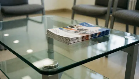 Une table basse en verre transparente posée sur le sol, portant plusieurs brochures de services. Des reflets lumineux sont visibles sur la surface. En arrière-plan flou, on aperçoit des chaises grises typiques d'un