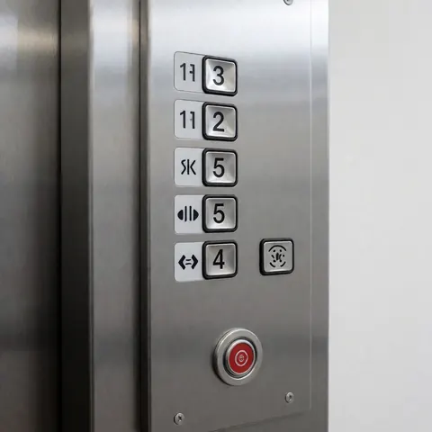 Panneau de contrôle d'ascenseur en acier inoxydable avec boutons pour les étages et bouton rouge d'urgence, monté sur un mur blanc sous un éclairage doux.