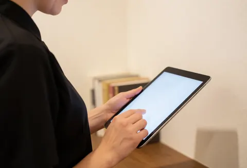 Un photographe professionnel examine son portfolio sur une tablette. Ses mains et son équipement sont visibles dans un bureau avec des livres flous en arrière-plan.