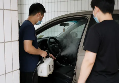 Un technicien portant un masque utilise une machine à vapeur pour désinfecter l'intérieur d'une voiture, avec le volant et les sièges visibles sur fond de murs carrelés blancs.