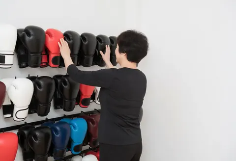 Une femme en tenue de sport touche des gants de boxe colorés posés sur une étagère dans un environnement lumineux et épuré.